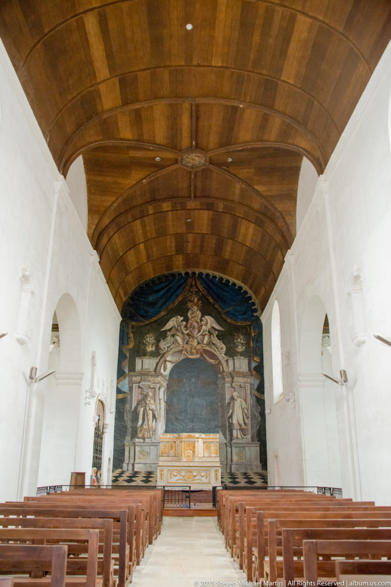 Whitewashed church interior with wooden ceiling, rows of pews, altar, and a large painted backdrop behind the sanctuary.
