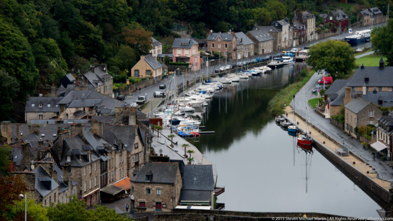 Elevated view of the Port of Dinan with sailboats moored along the River Rance between stone houses and a tree-lined quay.