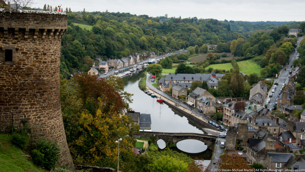 View from Dinan’s ramparts over the River Rance, port buildings, bridges, and wooded valley, with a stone tower edge at left.