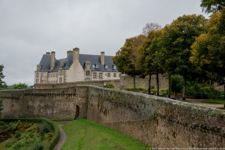 Curving stone rampart and grassy ditch in Dinan with a large historic slate-roofed residence and trees beyond.