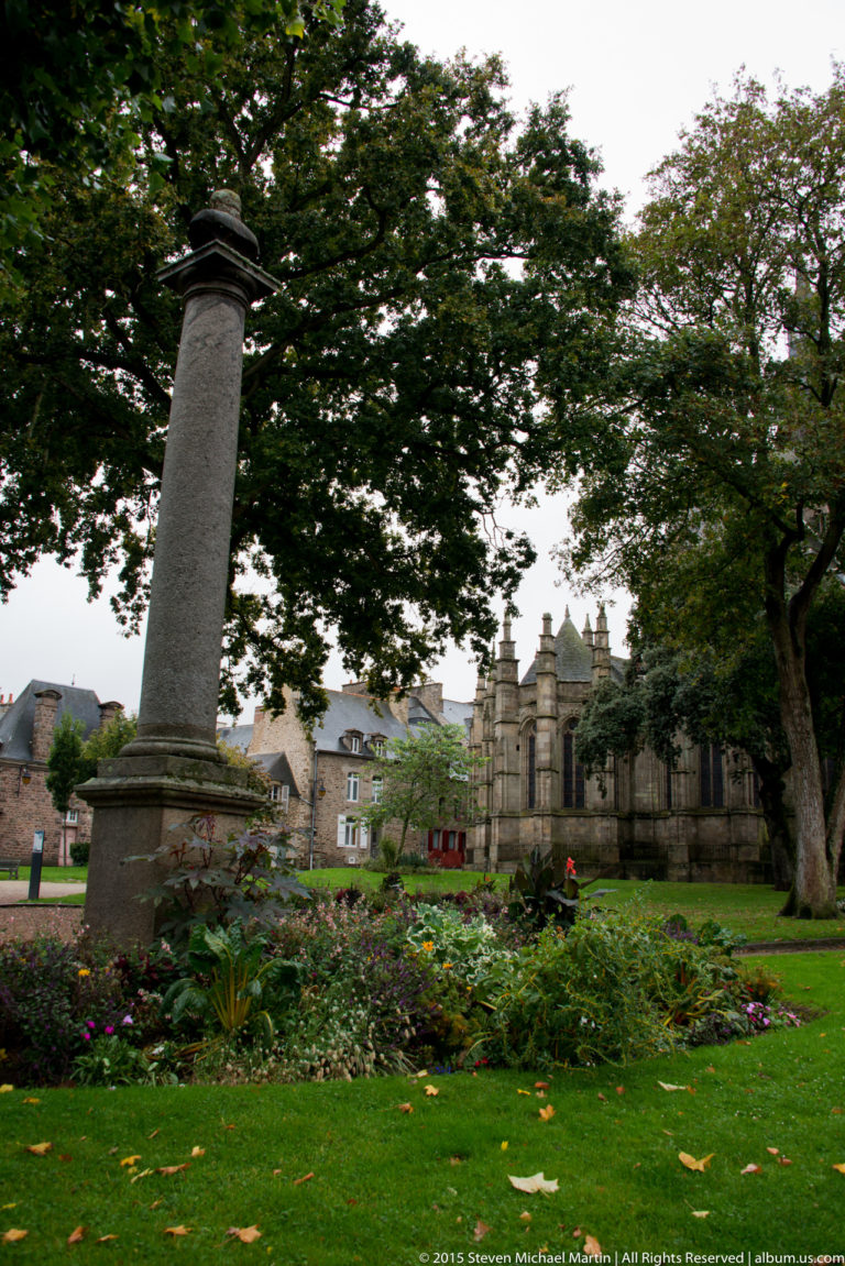 Tall plain stone column monument standing in a landscaped garden with mature trees, flowers, and historic buildings behind.
