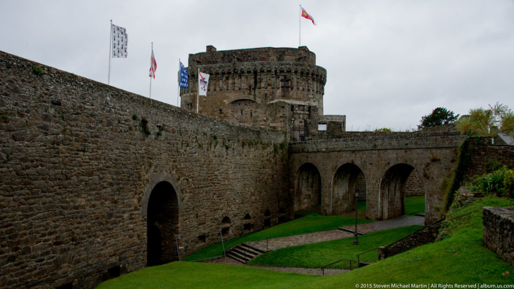 Massive stone walls, round tower, arched passages, grassy ditch, and flags at the Château de Dinan.