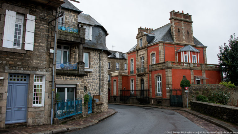 Curving Dinan street lined with stone and red stucco historic houses, slate roofs, balconies, and tall chimneys.