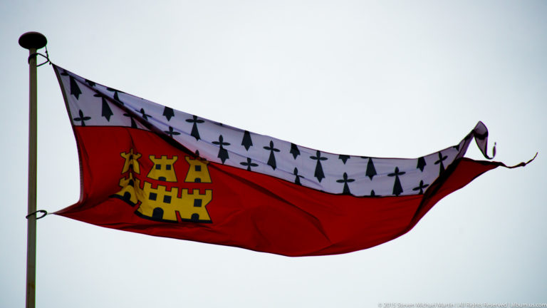 Red and white flag with black Breton ermine spots and a gold castle emblem flying against a pale sky.