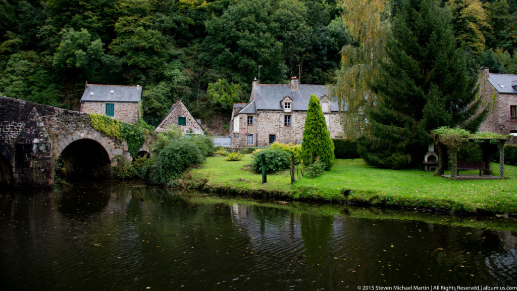Stone houses, gardens, and a low arched bridge beside calm water along the River Rance near Dinan.