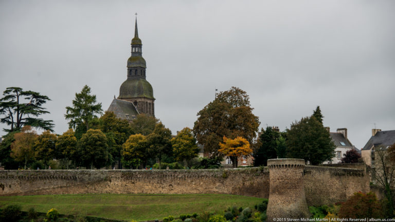 Church tower with a tall layered spire rising above autumn trees and stone walls in Dinan, Brittany.