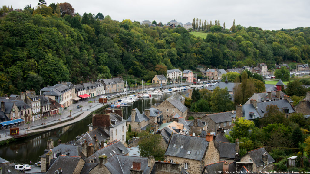 Elevated panorama of the Port of Dinan with moored boats, River Rance, slate-roofed houses, and wooded hillsides.
