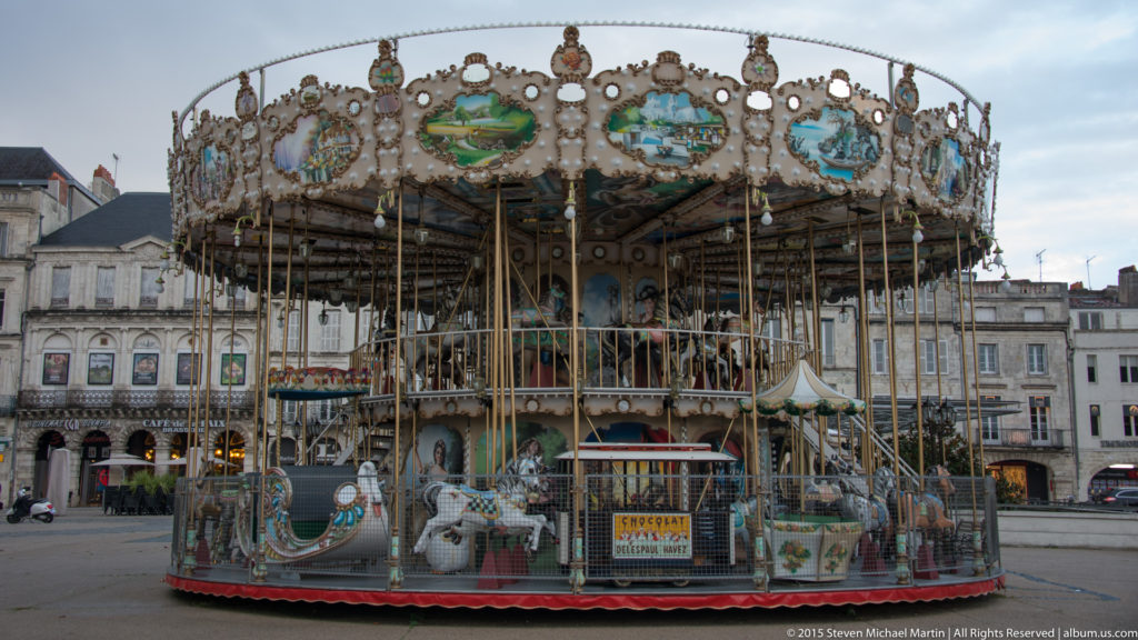 Ornate traditional carousel with painted panels and ride figures standing in a paved square among pale stone buildings in La Rochelle.