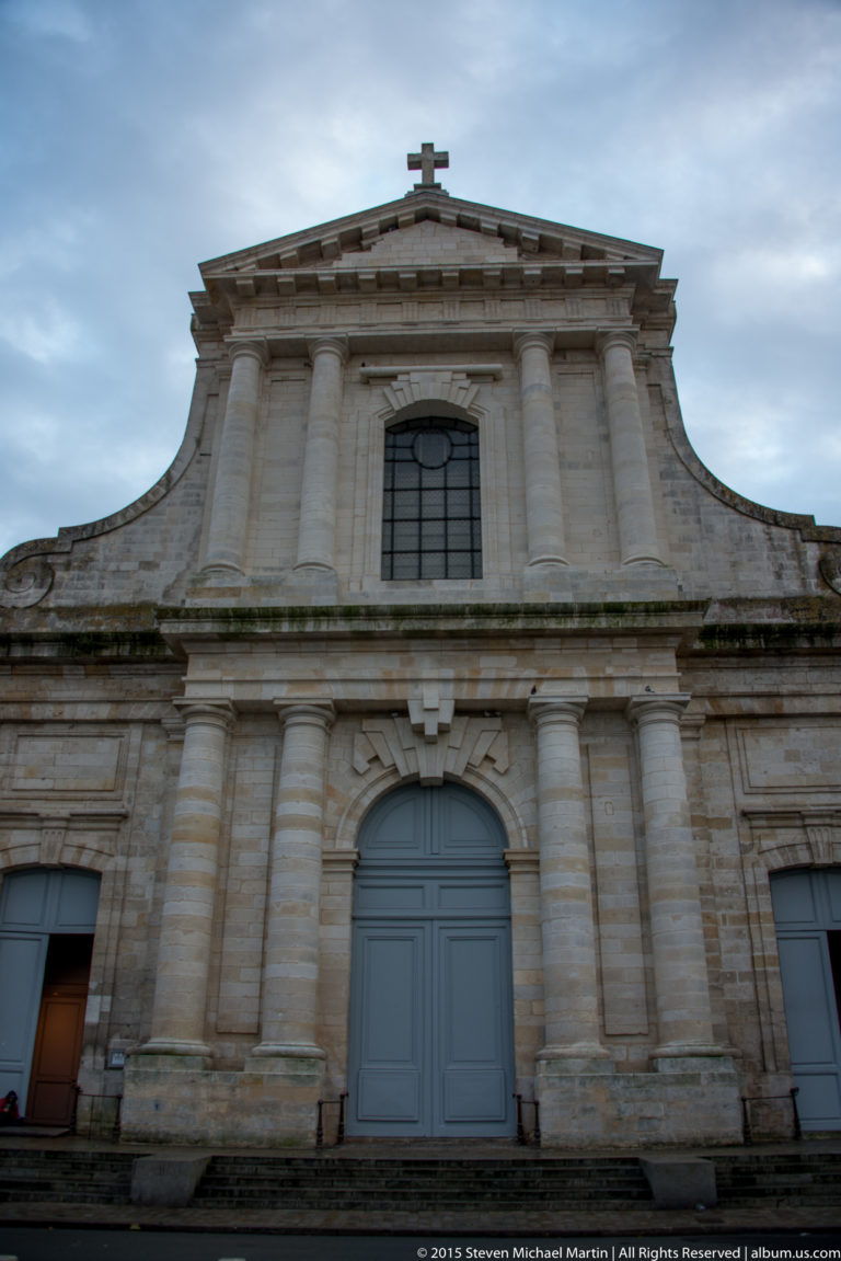 Symmetrical pale-stone church façade with arched central doorway, pilasters, curved upper profile, and a cross at the roofline.