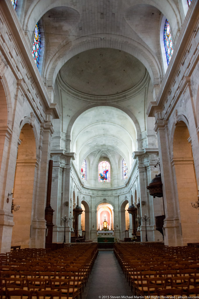 Wide church nave with pale stone arches, vaulted ceiling, wooden seating, stained-glass windows, and an altar centered in the distance.
