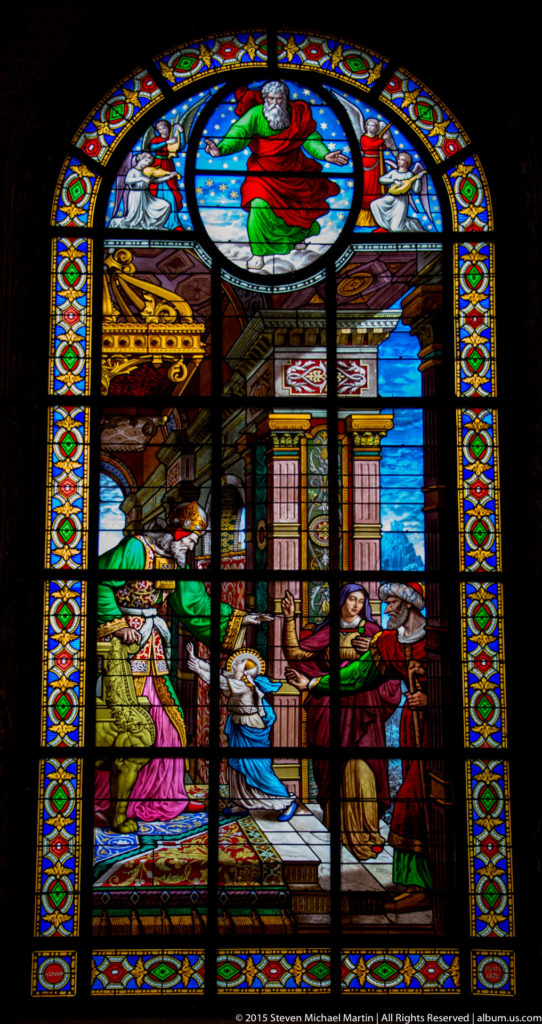 Tall multicolored stained-glass window with a Christian narrative scene, figures, architectural motifs, and an upper circular medallion.