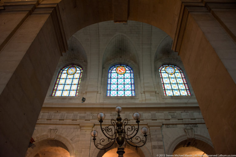 Three illuminated stained-glass windows above a large dark metal chandelier, framed by pale stone arches inside a church.