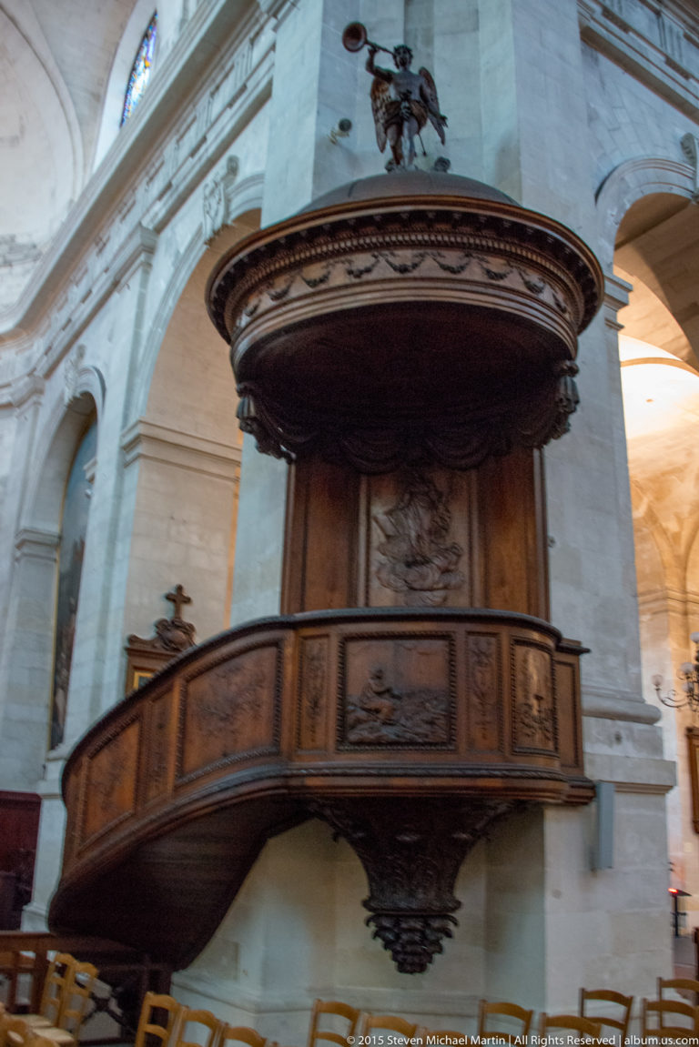 Carved dark wooden pulpit projecting from a pale stone pier, with paneled sides and a broad circular sounding board above.
