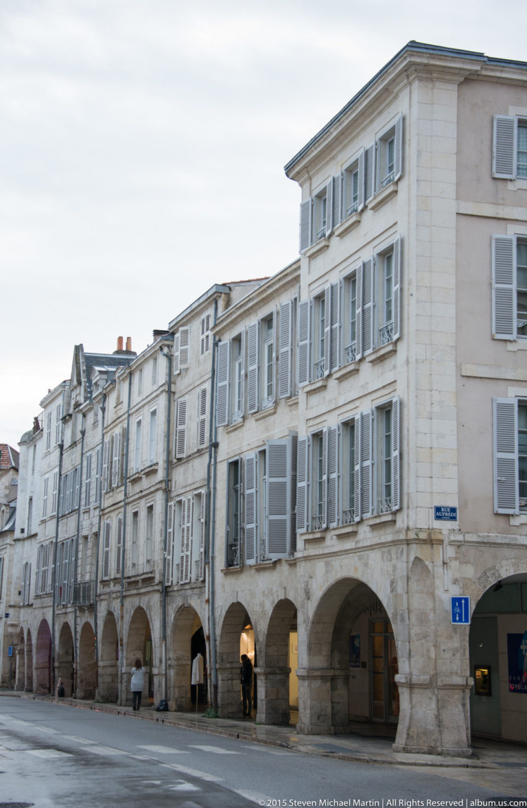 Historic pale façades with shuttered windows and continuous ground-floor arcades lining a street in central La Rochelle.