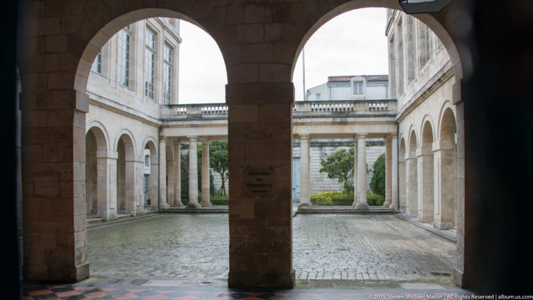 View through a dark stone arch into a pale cobbled courtyard with arcades, columns, and an upper balustrade.