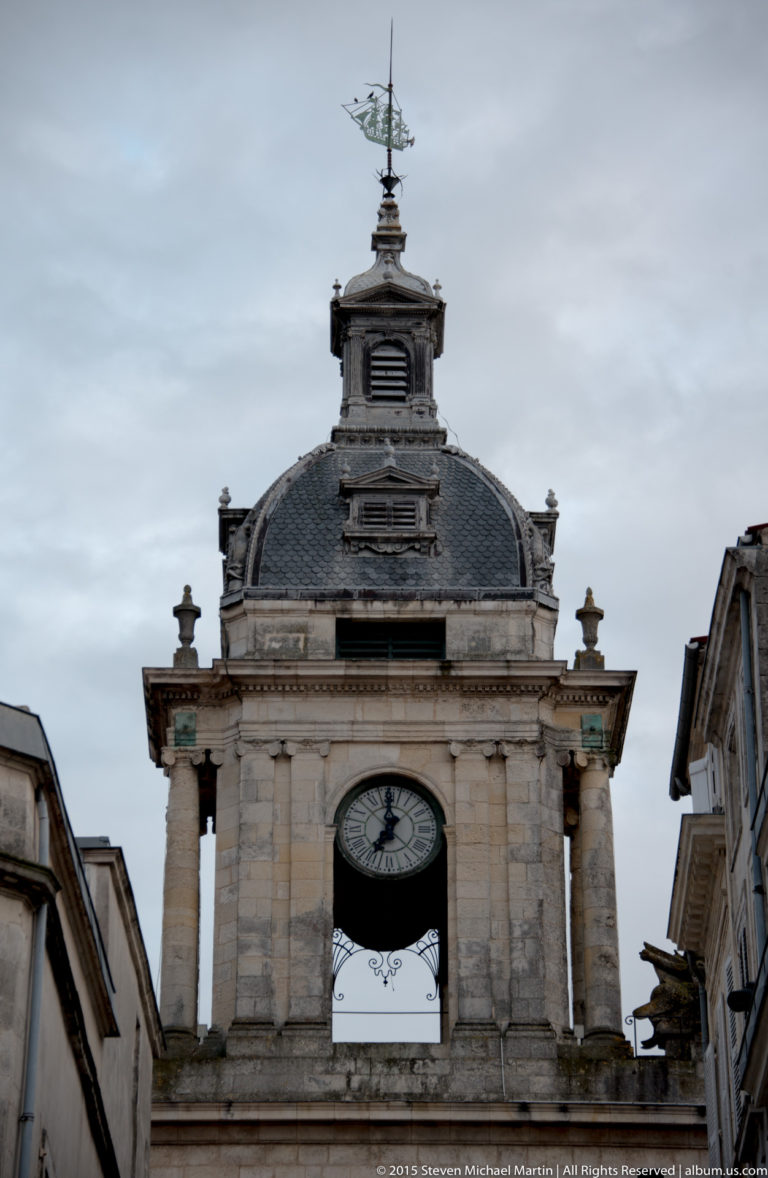 Upper Grosse Horloge in La Rochelle with large clock face, ornate slate-roofed pavilion, belfry, finials, and weather vane.