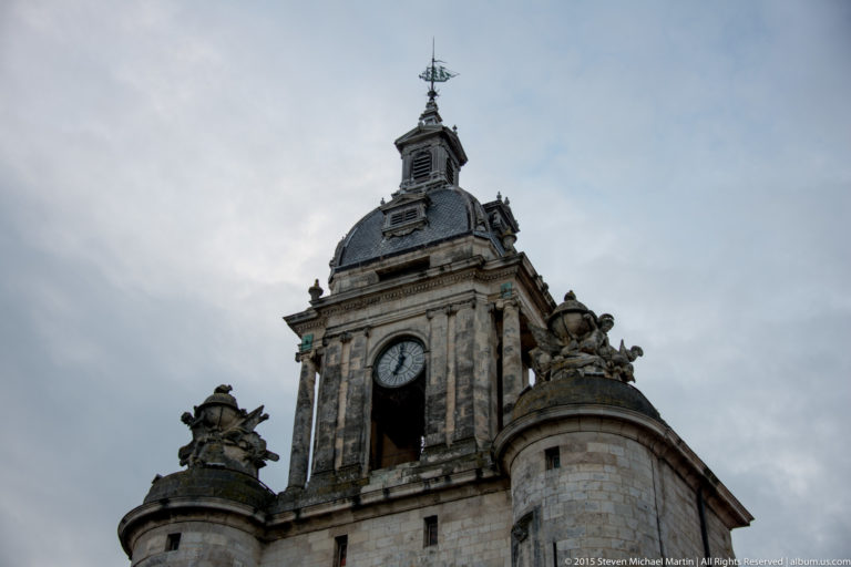 Low-angle oblique view of the Grosse Horloge with masonry clock tower, slate pavilion, finials, and sculptural roofline.