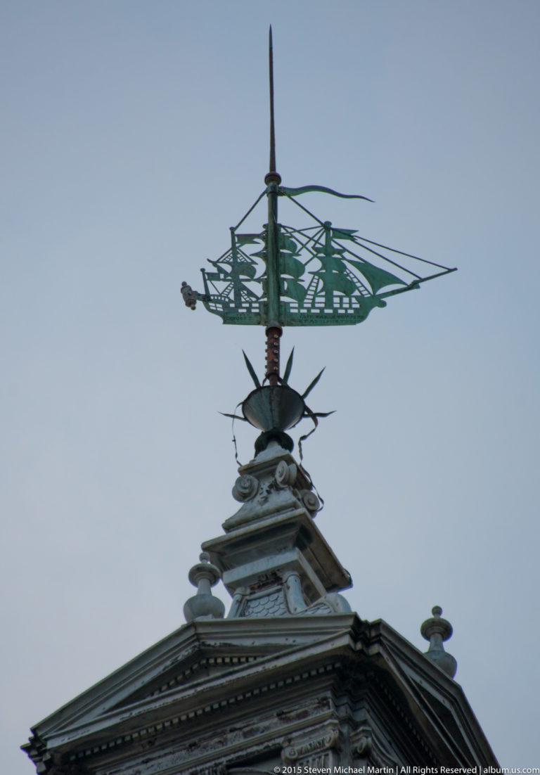 Tight silhouette of the Grosse Horloge’s ship-shaped metal weather vane and roof finial against a gray sky.