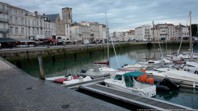 Small boats moored in La Rochelle’s old harbor with masts, calm water, and a long row of pale waterfront buildings.