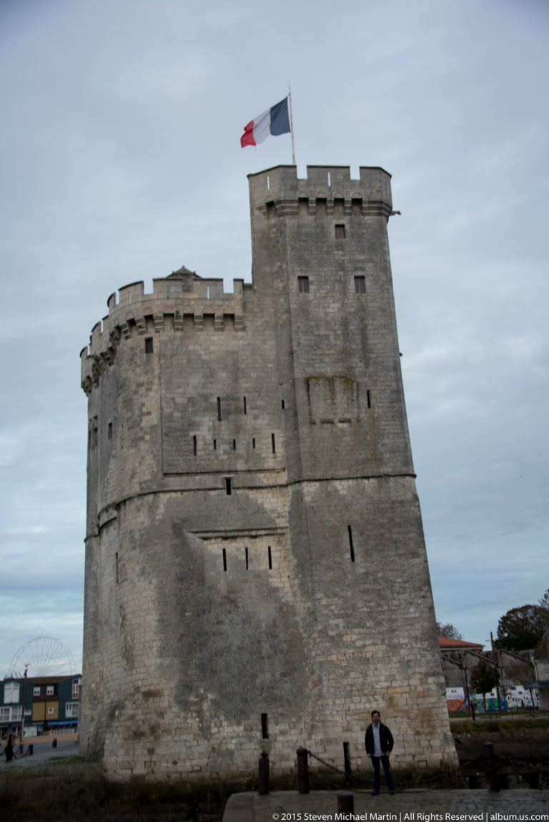 Close frontal view of crenellated Tour Saint-Nicolas with French flag, narrow openings, and a person near the base for scale.