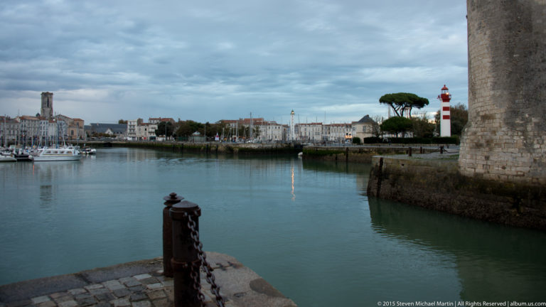 Calm La Rochelle harbor with waterfront buildings and red-and-white harbor light, framed at right by the stone wall of a tower.