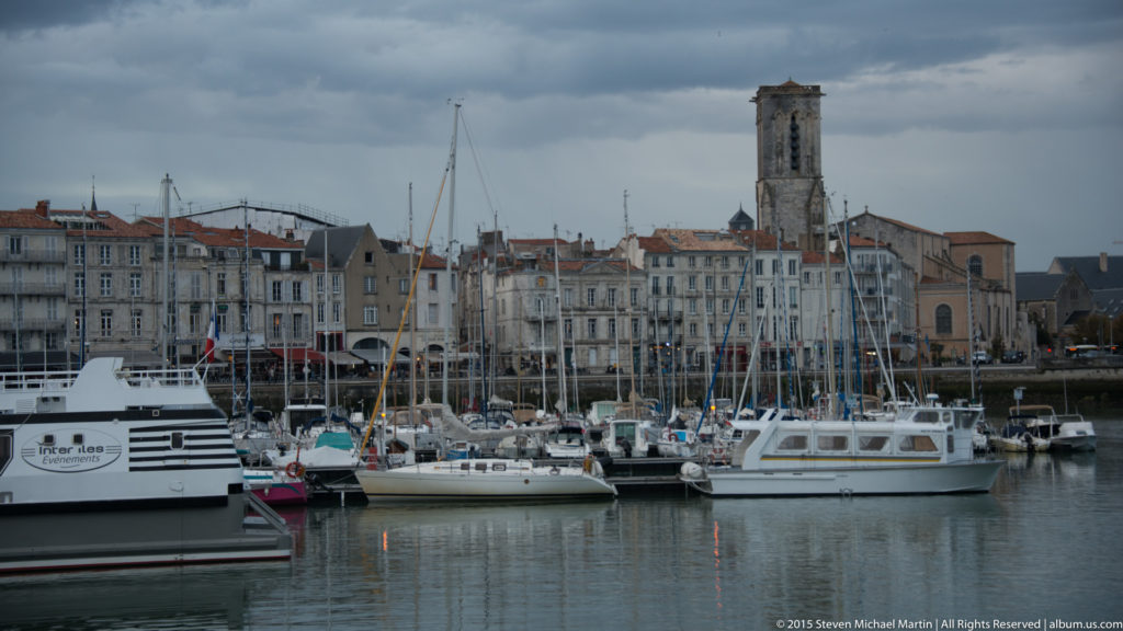 Crowded marina in La Rochelle’s old harbor with sailboats, motor boats, waterfront buildings, and a distant stone church tower.