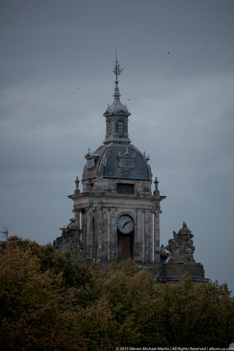 Distant upper Grosse Horloge visible above trees, with clock opening, ornate slate roof, finials, and ship weather vane.