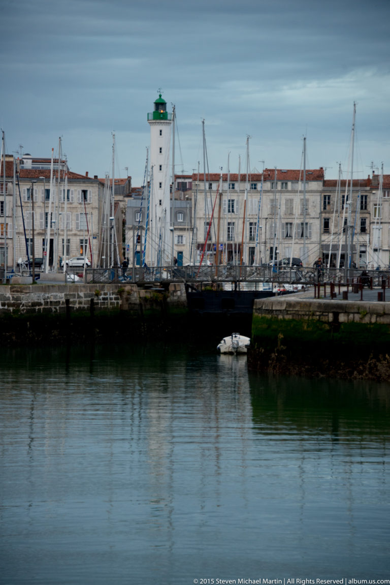Small white lighthouse with green lantern beside La Rochelle’s old harbor, backed by sailboat masts and waterfront buildings.