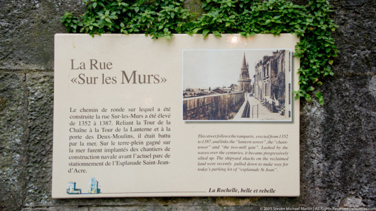 Bilingual plaque titled La Rue «Sur les Murs» with historical photograph and explanatory text mounted on a stone wall beside ivy.