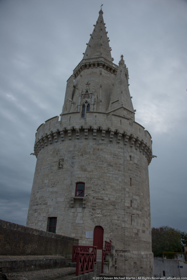 Close low-angle view of Tour de la Lanterne showing rounded masonry, crenellated gallery, small Gothic openings, and steep stone spire.