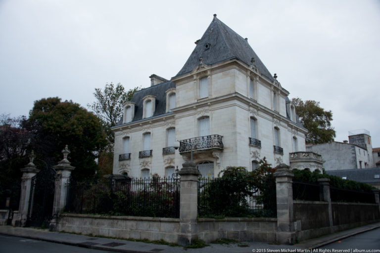 Large pale-stone historic residence with steep slate roof, dormers, balcony, garden, and iron fence along a La Rochelle street.