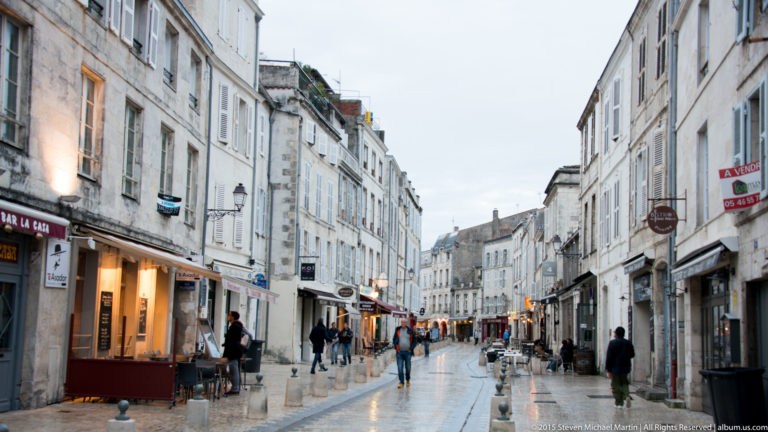 Narrow wet street in central La Rochelle lined with pale historic buildings, shops, cafés, bollards, and pedestrians.