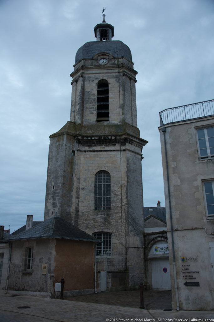 Square stone tower with louvered upper openings, small circular clock or medallion, low roof, and weather vane in La Rochelle.