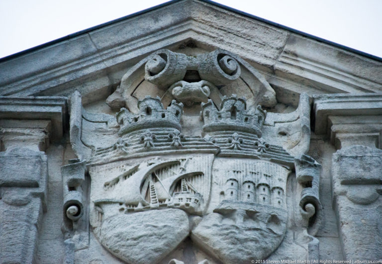 Weathered carved stone coat of arms with crowns, scrolls, a sailing vessel, and fortified architectural motifs.