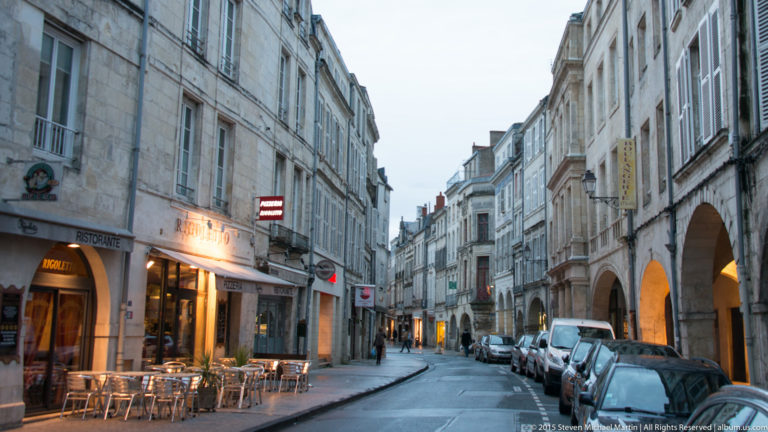 Narrow La Rochelle street lined with pale historic façades, shops, café tables, parked cars, and deep stone arcades at right.