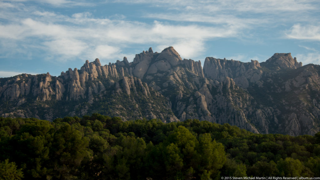 Jagged pale peaks of Montserrat rise above forested slopes beneath broken blue-and-white clouds.