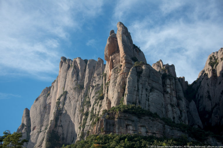 Steep weathered rock pinnacles of Montserrat rise against blue sky, with trees and shrubs at their base.