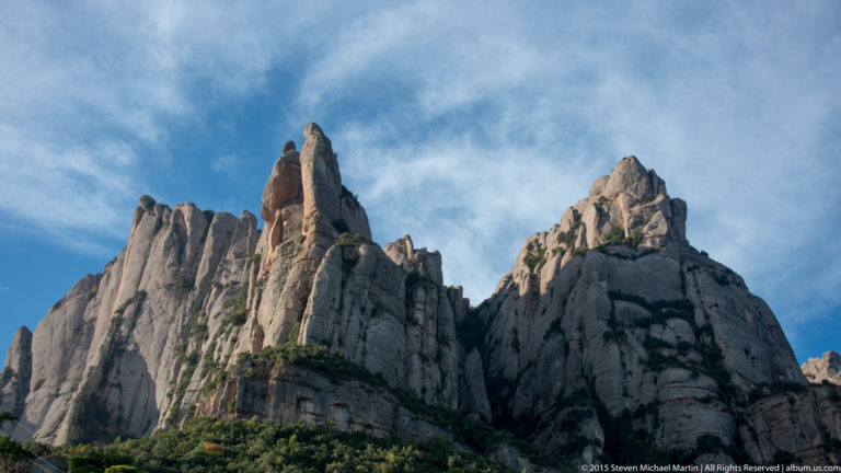 Clustered gray rock towers of Montserrat rise steeply above green ledges under a blue, cloud-streaked sky.