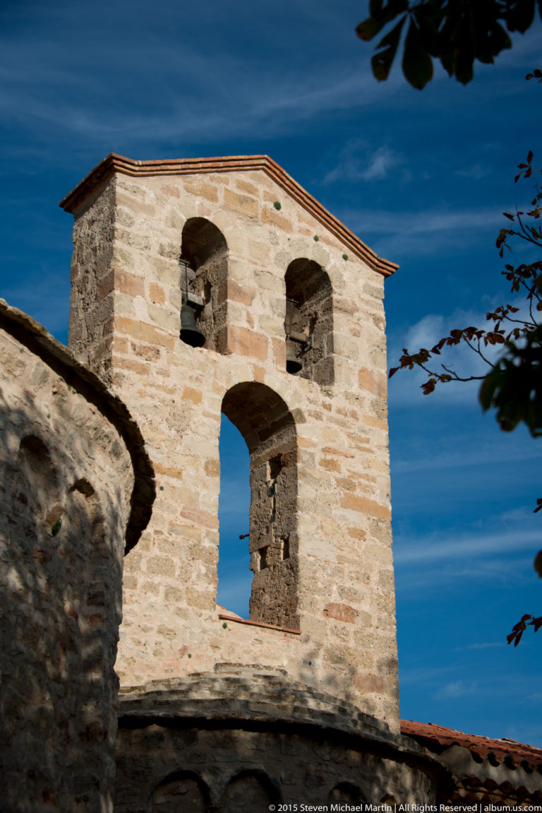 Stone bell gable with three arched openings and tiled cap, framed by dark foliage against blue sky.
