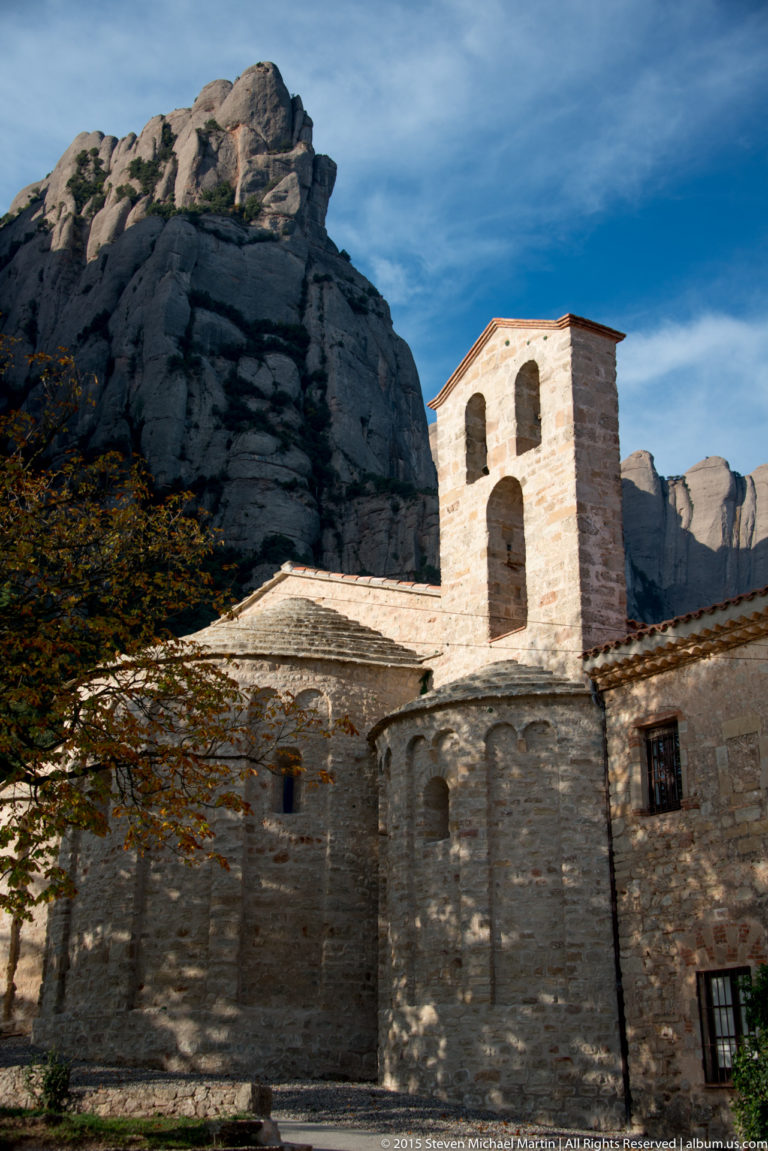 Small stone church with bell gable and rounded apse stands beneath the massive cliffs of Montserrat.