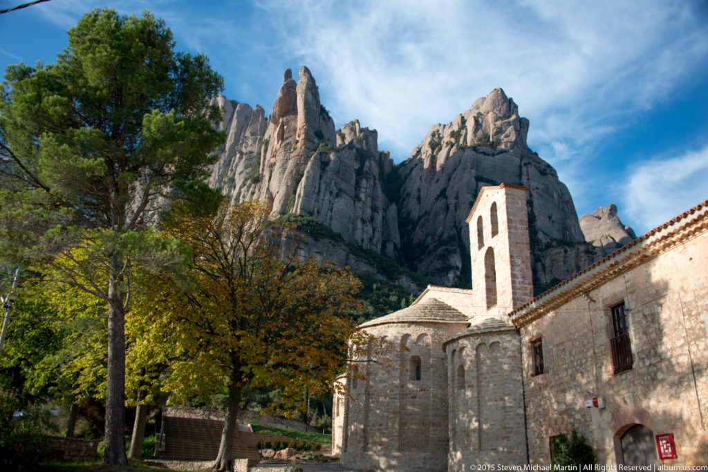 Stone church complex among trees, with bell gable and Montserrat rock pinnacles rising behind.