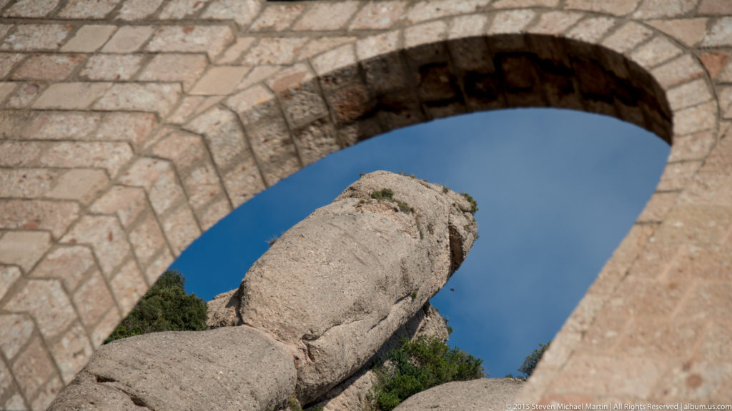 Rounded Montserrat rock pinnacle seen through a large weathered stone arch against deep blue sky.