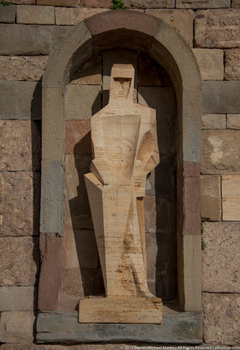 Angular stone figure in a shallow arched wall niche, lit strongly from the side.