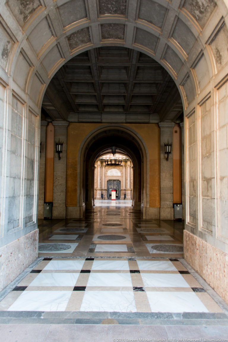 Long arched monastery corridor with checkered floor, paneled ceiling, and strong central perspective.