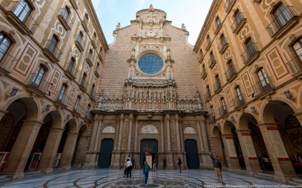 Ornate stone façade of the Montserrat basilica with central rose window, sculpture, and flanking arcaded buildings.