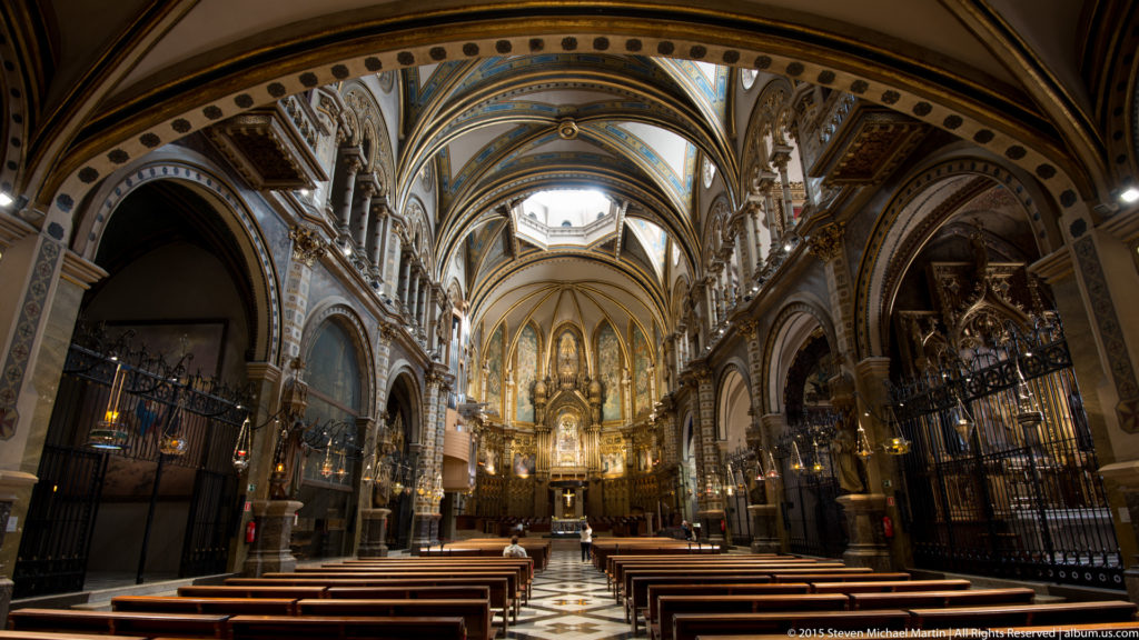 Central nave of Montserrat basilica with wooden pews, decorated arches, iron screens, and gilded sanctuary.