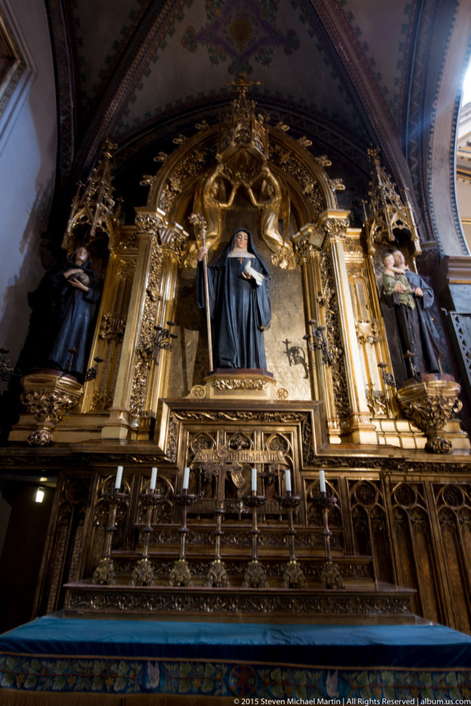 Black-robed saint centered in an ornate gilded side altar with flanking figures inside Montserrat basilica.