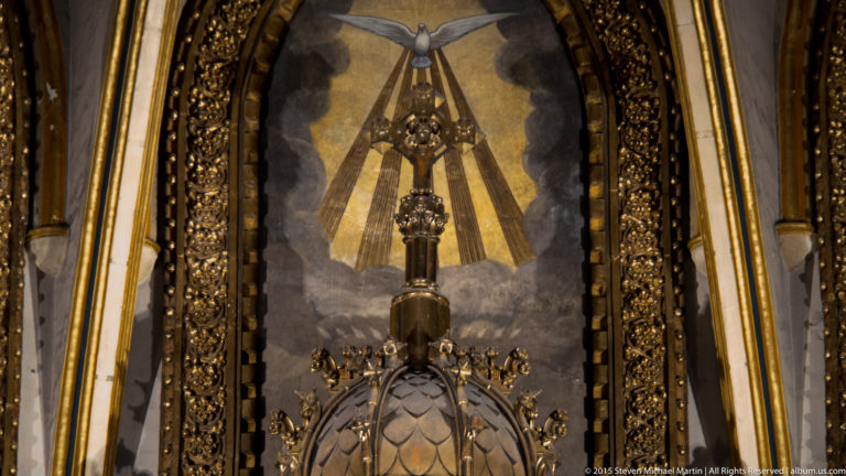 Ornate dark hanging lamp centered before a pale Marian devotional image with radiating triangular forms.