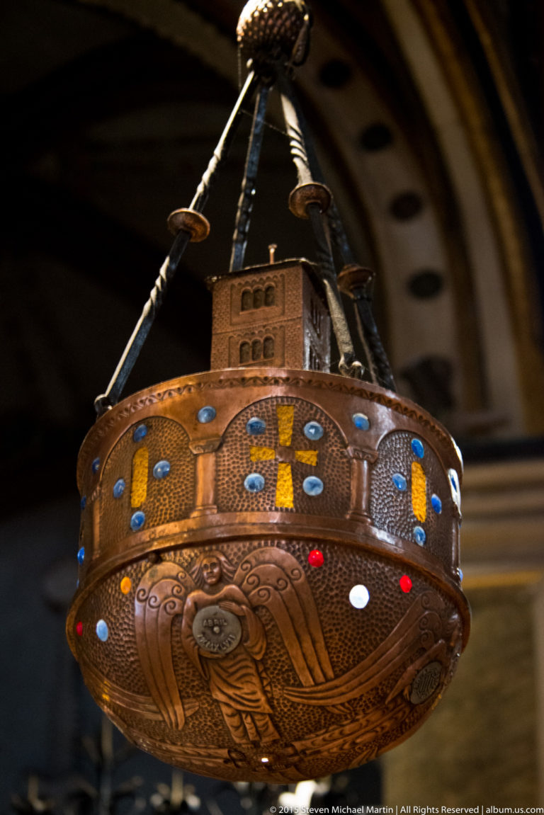 Copper-toned hanging lamp with sacred figure, colored crosses, blue and red insets, and suspension straps.