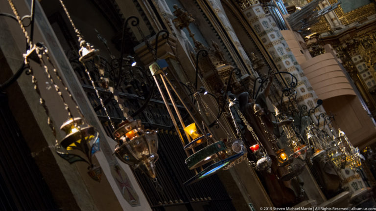 Rows of ornate hanging lamps and chains recede through the decorated interior of Montserrat basilica.